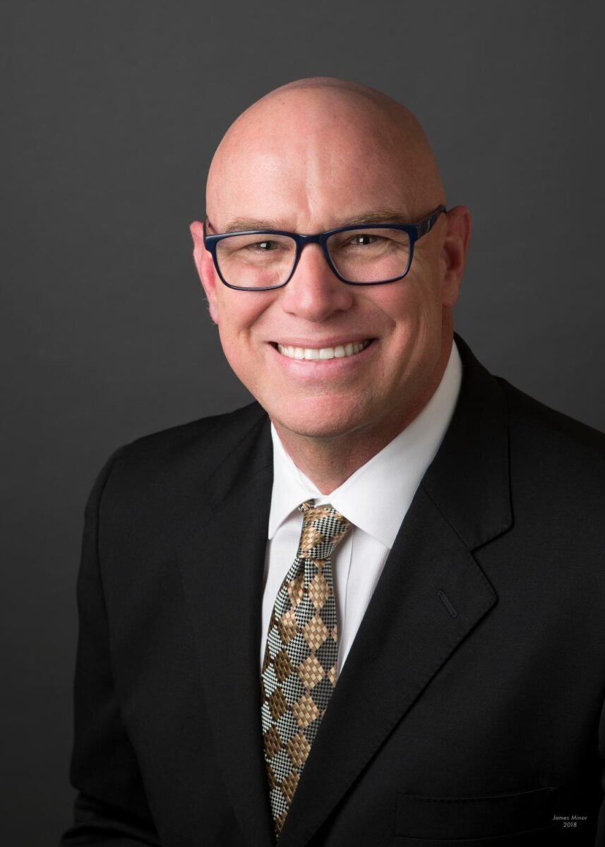 Alt text: "Portrait of a smiling bald man with blue-framed eyeglasses. He is wearing a black suit, white shirt, and a patterned tie with brown, tan, and blue squares. The background is a neutral dark gray, providing a professional appearance. The man's confident and cheerful expression suggests a friendly and approachable personality. The photo is marked 'James Minor 2018' in the bottom right corner, indicating the photographer's name and the year the photo was taken.