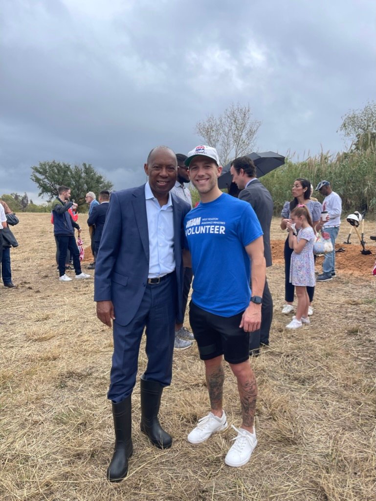 A photo of two men standing side by side, smiling at the camera in an outdoor setting with a cloudy sky overhead. On the left is a man in a dark suit and rubber boots, and on the right is a younger man in a blue 'TEAM VOLUNTEER' t-shirt, black shorts, and white sneakers, with visible tattoos on his legs. Around them are other people in a casual outdoor gathering, some with umbrellas, suggesting a community event or volunteer activity.