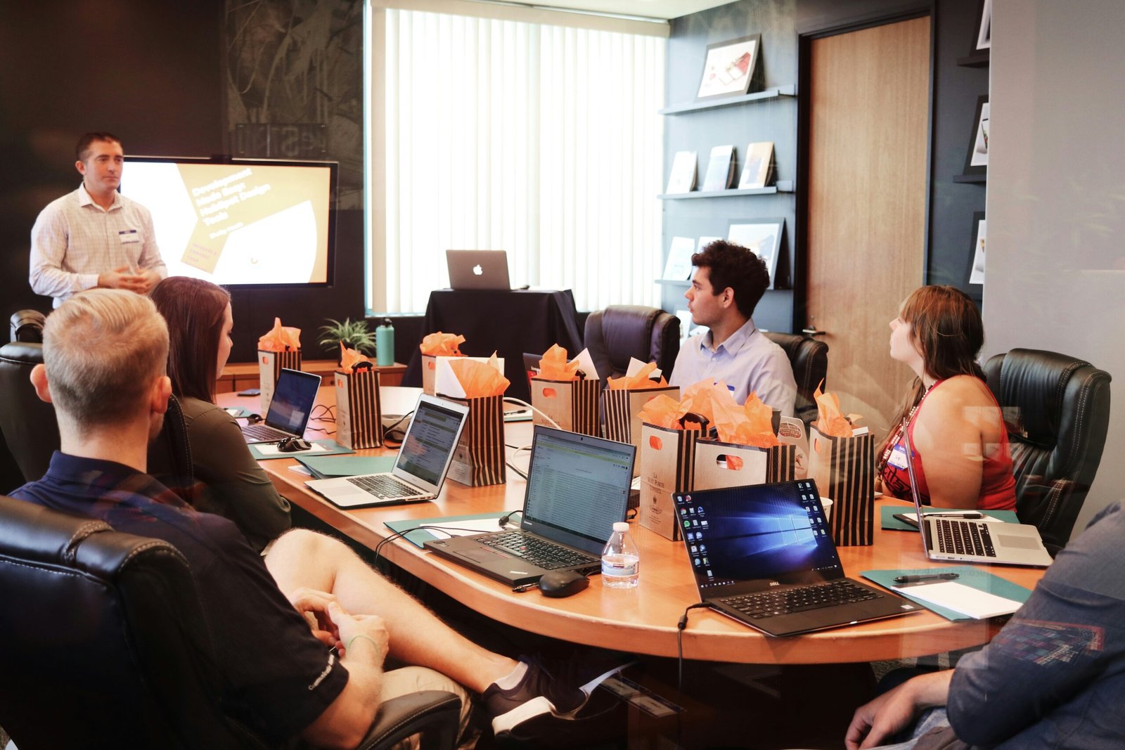 This image shows a professional setting where a presentation is taking place. There is a person standing at the front, presumably leading the session, with a screen behind them showing what looks like a slide from a presentation. The audience, consisting of several individuals, is seated around a conference table. Most are facing the presenter, indicating attention and engagement. They have laptops open in front of them, suggesting that this is a work-related meeting where note-taking or immediate follow-up on the discussion points might be needed. The room appears well-lit, and there are gift bags on the table, which could imply that this event is special, perhaps a seminar or a workshop with some token of appreciation provided to the attendees.